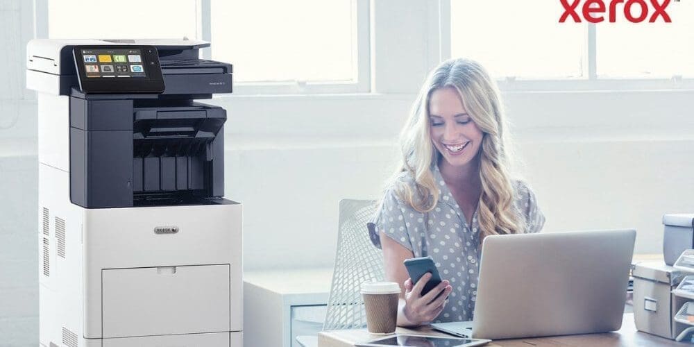 Eine Person schaut zufrieden auf ihr Smartphone, während sie mit einem Laptop an einem Schreibtisch sitzt. Neben dem Schreibtisch in einem hell erleuchteten Büroraum ist ein Multifunktionsdrucker Xerox VersaLink B605 zu sehen.