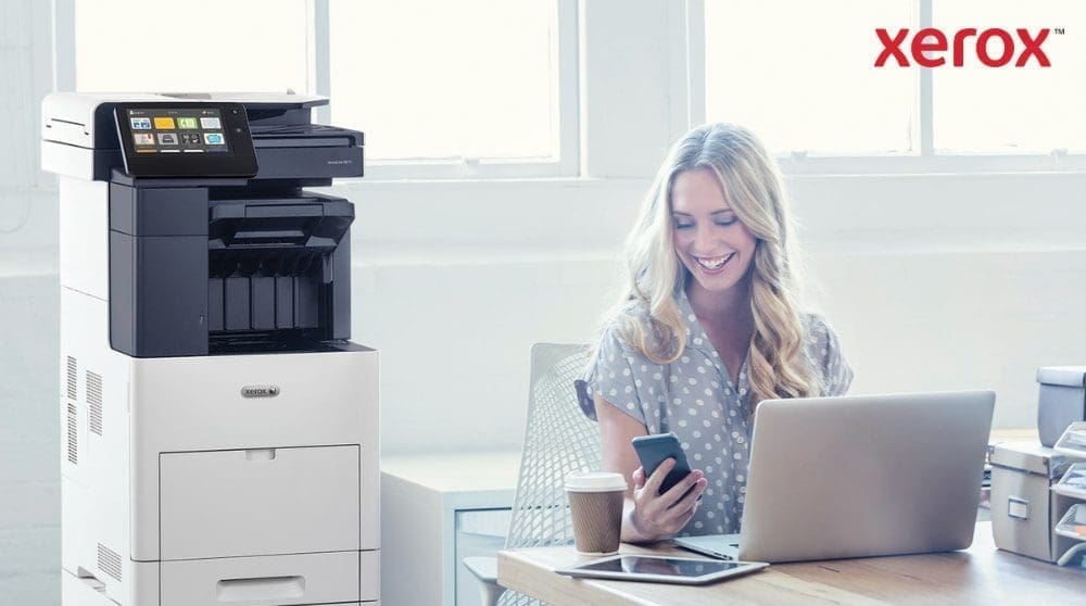 Eine Person schaut zufrieden auf ihr Smartphone, während sie mit einem Laptop an einem Schreibtisch sitzt. Neben dem Schreibtisch in einem hell erleuchteten Büroraum ist ein Multifunktionsdrucker Xerox VersaLink B605 zu sehen.