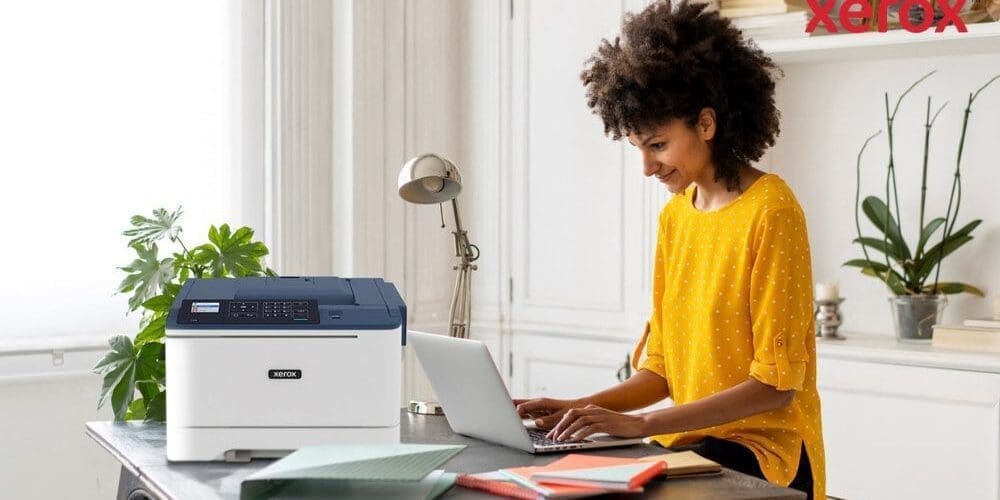 Eine Person mit lockigem Haar lächelt, während sie an einem Schreibtisch auf einem Laptop tippt. Neben dem Laptop steht ein kompakter Xerox C310-Drucker, der ein modernes Home-Office-Setup für mehr Produktivität vorführt.