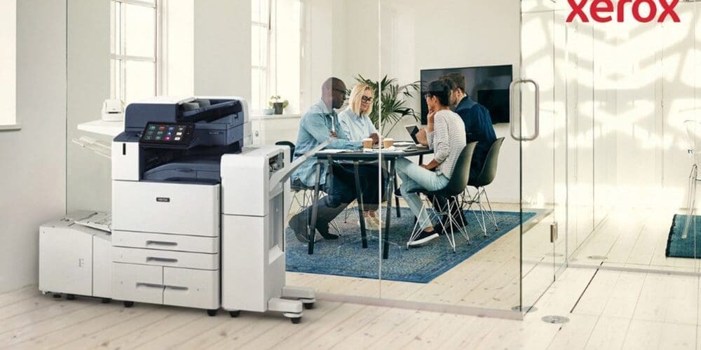 Im Vordergrund eines hellen Büros steht ein moderner Multifunktionsdrucker Xerox AltaLink B8245. Im Hintergrund sitzt eine Gruppe von Menschen an einem Tisch und diskutiert gemeinsam.