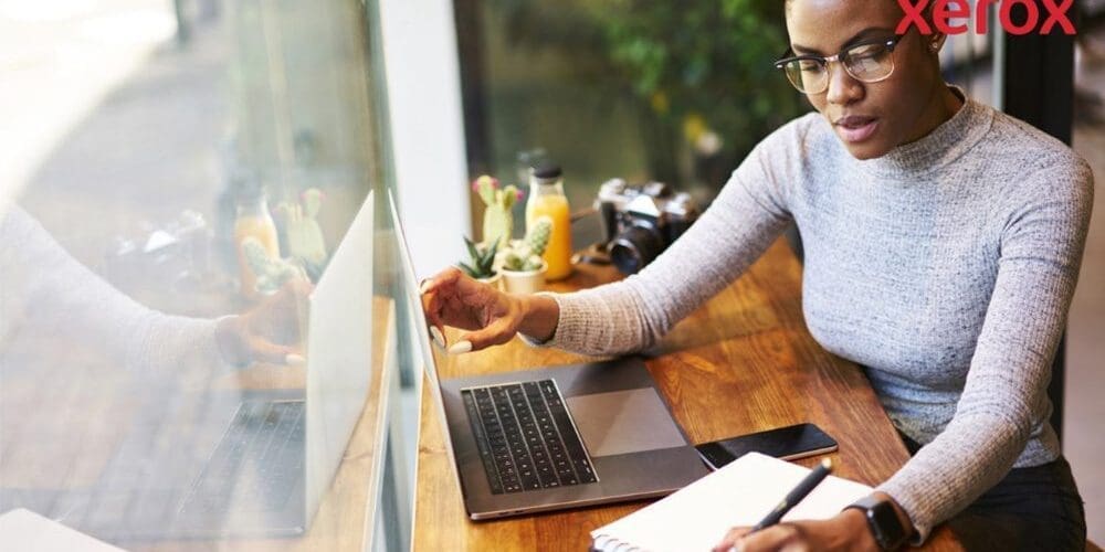 Eine Person mit Brille arbeitet an einem Holztisch, schaut auf einen Laptop-Bildschirm und schreibt in ein Notizbuch, während ein Smartphone in der Nähe ist, was auf eine produktive Arbeitsumgebung hindeutet